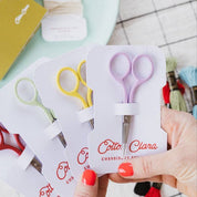 Hand holding a set of small scissors in packaging with 'Cotta Clara' branding on a checkered table.