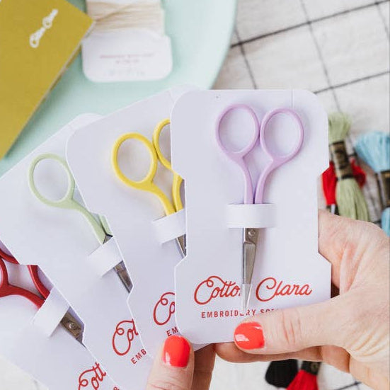 Hand holding a set of small scissors in packaging with 'Cotta Clara' branding on a checkered table.