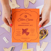 Cotton Clara wooden birds embroidery kit on a purple background with craft supplies.