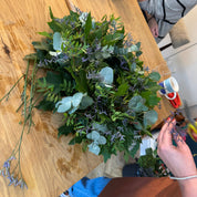 Person arranging greenery on a wooden table with another person partially visible.
