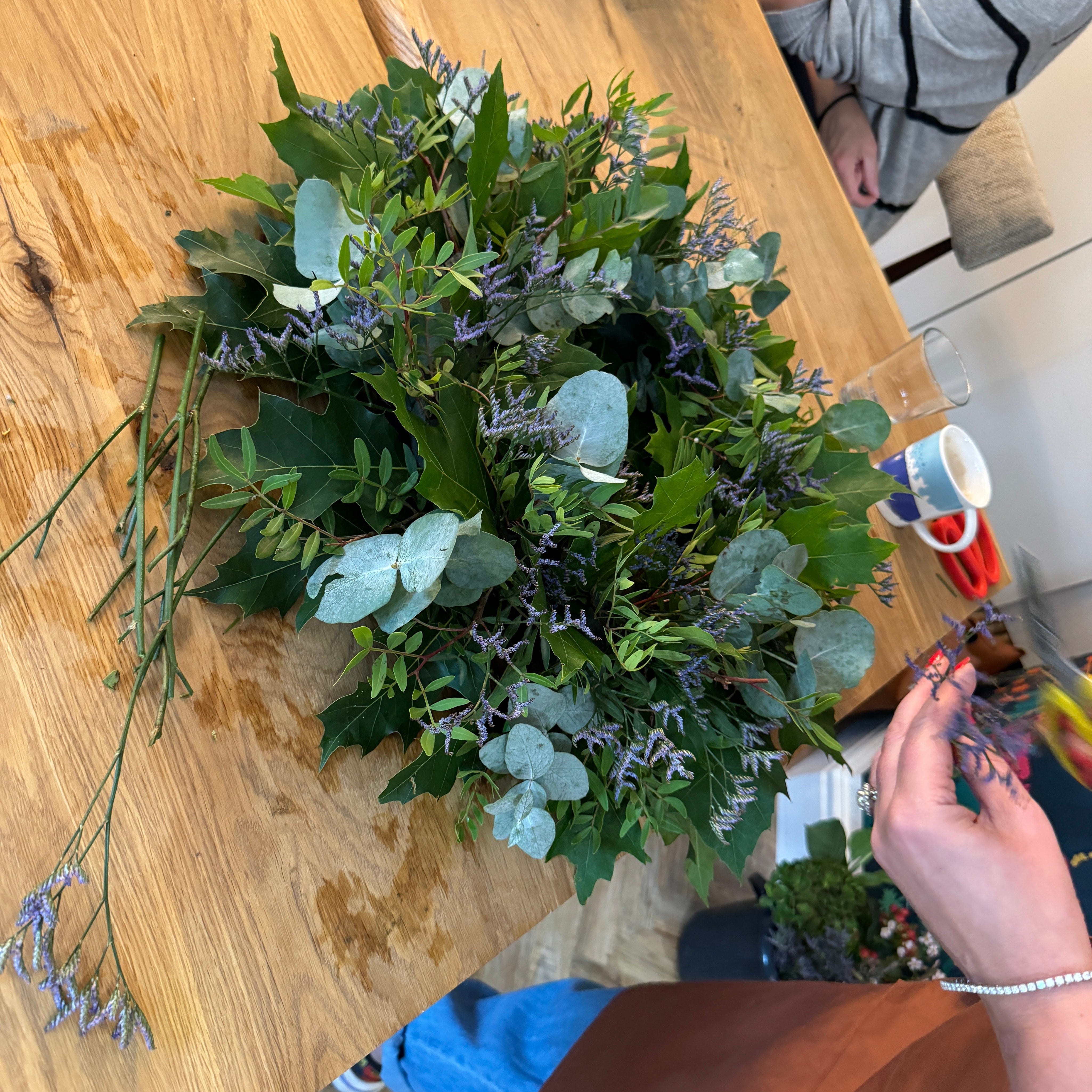 Person arranging greenery on a wooden table with another person partially visible.