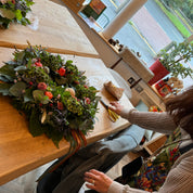 Person arranging flowers on a table in a room with large windows.