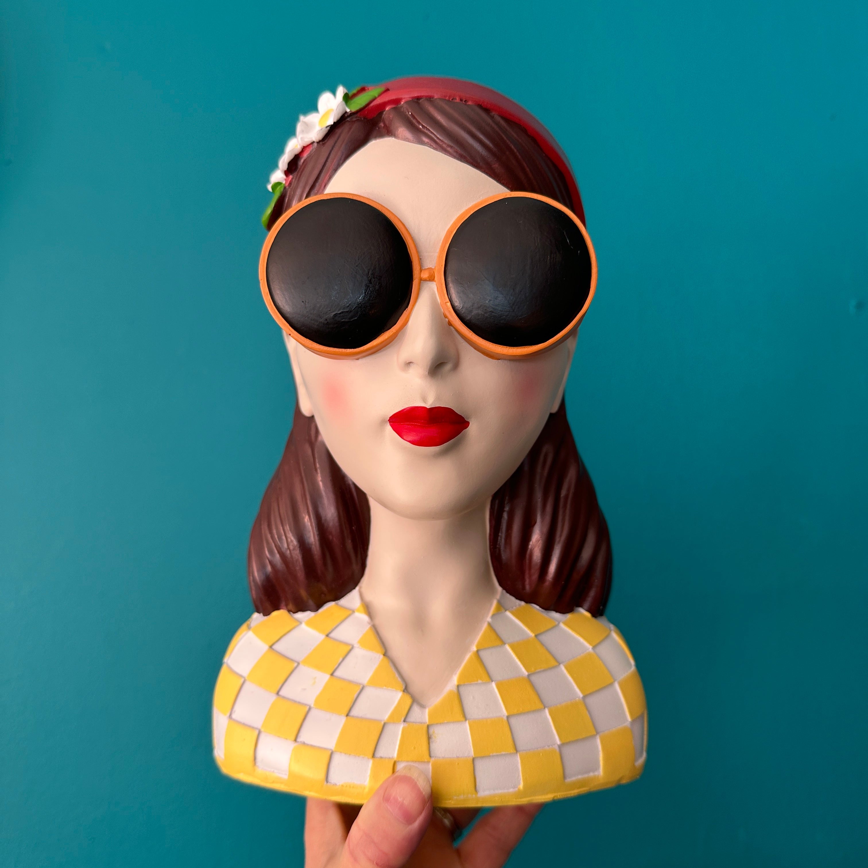 Decorative head with sunglasses and a yellow checkered hat held against a teal background