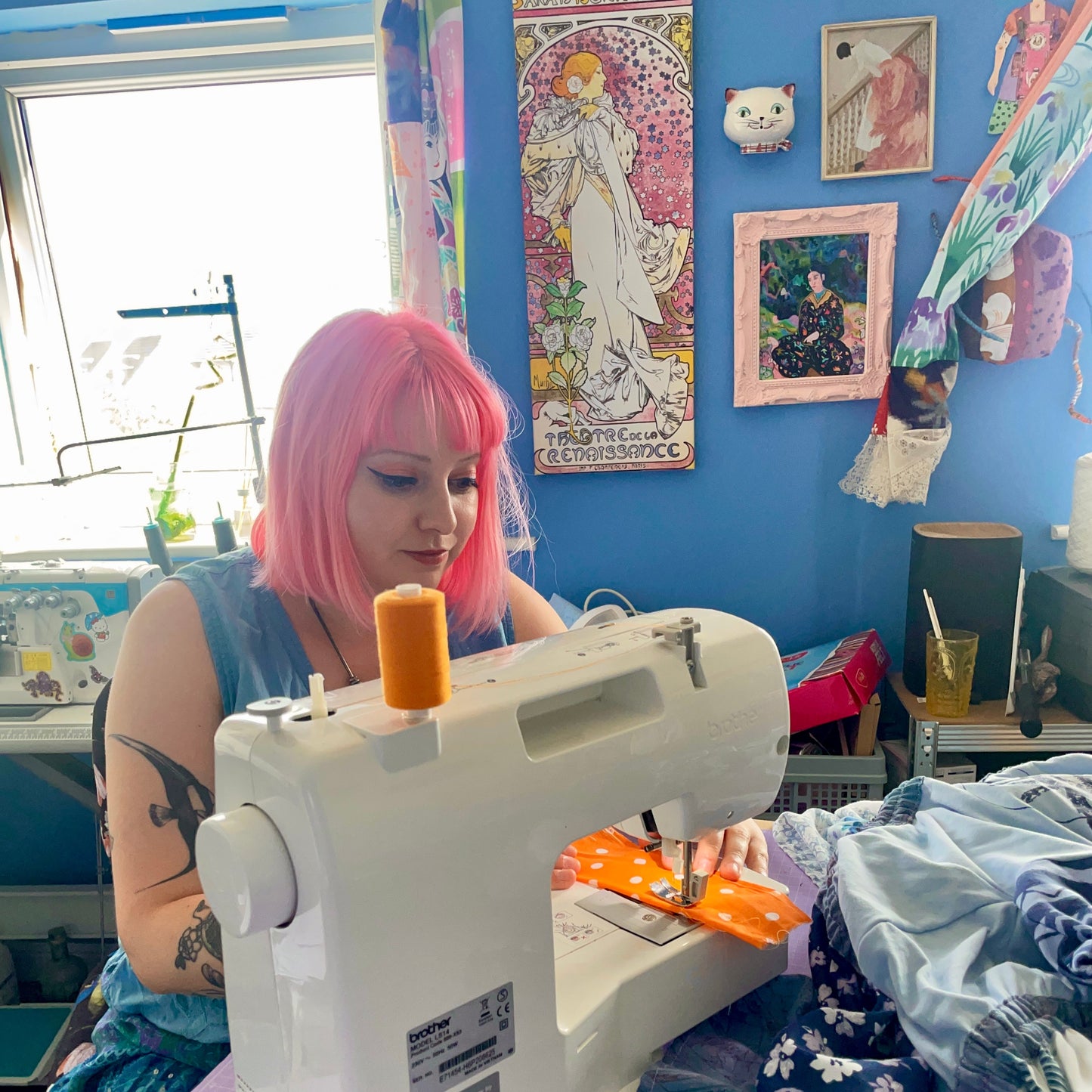 Person with pink hair using a sewing machine in a room with blue walls and various decorations.
