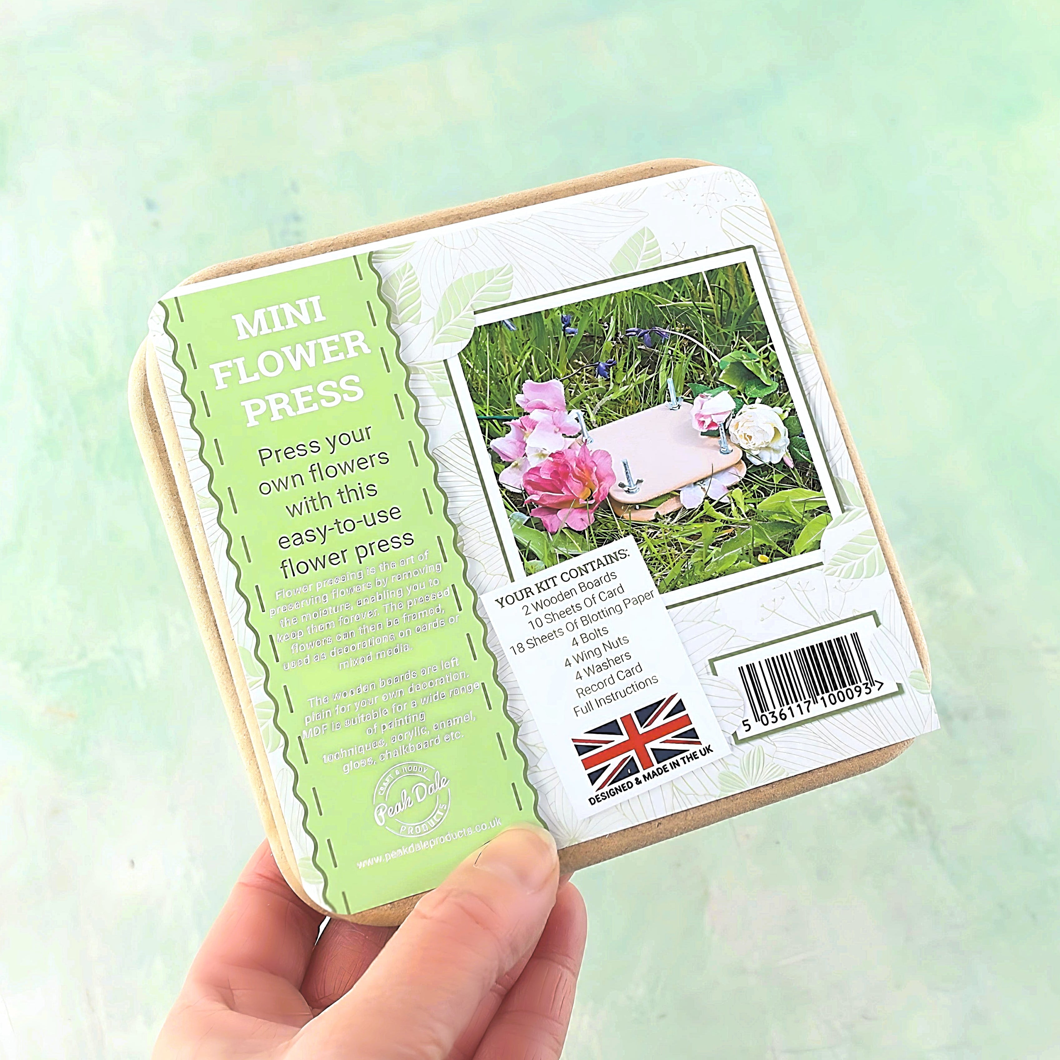 A hand holding a Mini Flower Press with its packaging visible. The packaging has an image of pressed flowers and text describing the product.