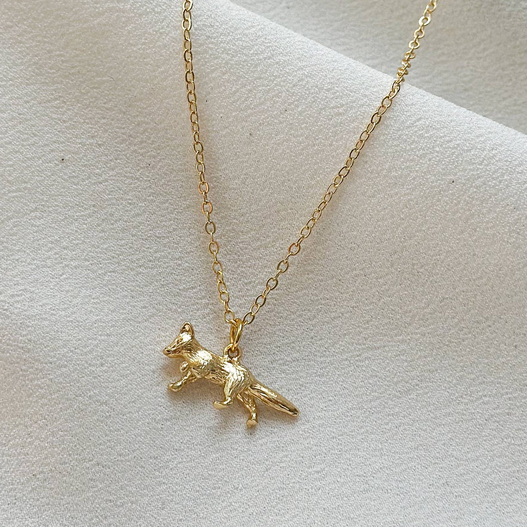 A delicate gold chain necklace featuring a small, textured gold dog pendant, displayed on a soft, draped white fabric background.