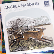 Angela Harding 2027 Calendar featuring a detailed woodcut-style illustration of a hare, set against a stack of other colorful calendars on a light blue surface.