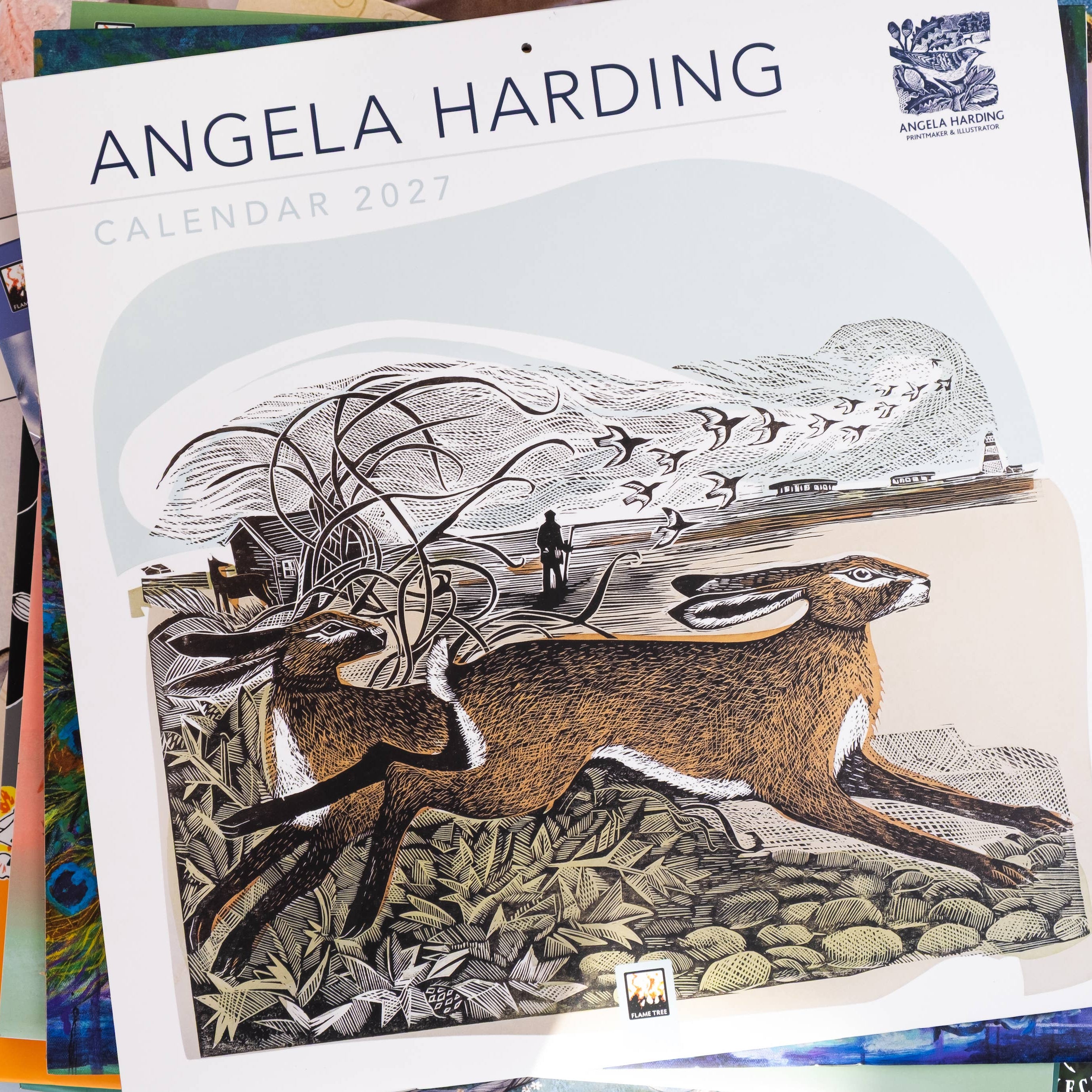 Angela Harding 2027 Calendar featuring a detailed woodcut-style illustration of a hare, set against a stack of other colorful calendars on a light blue surface.