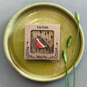 A small enamel pin by Lily Faith featuring a bird with a red chest and brown wings, mounted on a kraft paper card. The card is placed on a round, speckled olive-green ceramic plate, with two green poppy pods on the right side, all set against a neutral gray background.