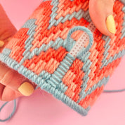 A close-up of a hand holding a small knitted or crocheted fabric item with a coral-orange and light blue geometric pattern, against a solid pink background.