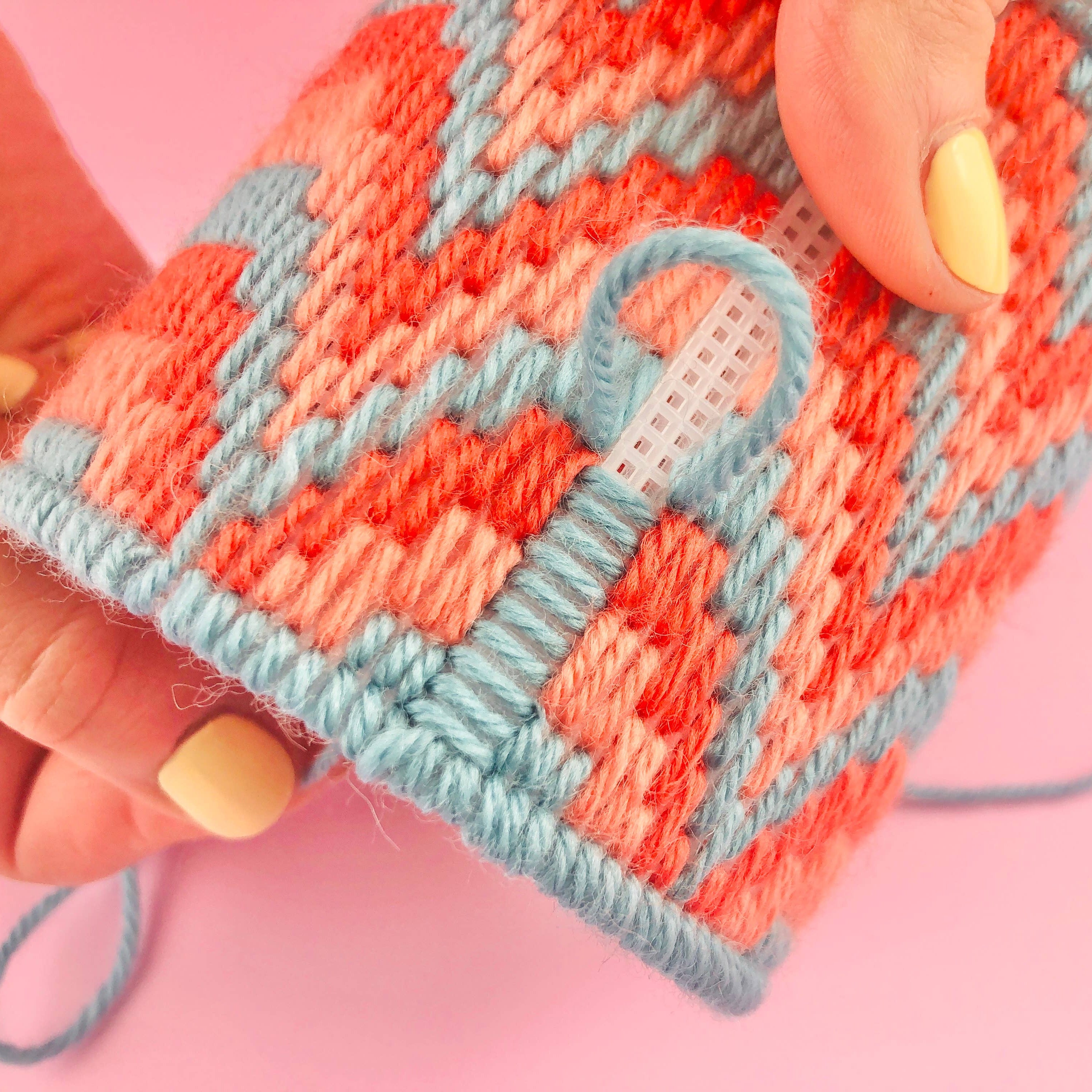 A close-up of a hand holding a small knitted or crocheted fabric item with a coral-orange and light blue geometric pattern, against a solid pink background.