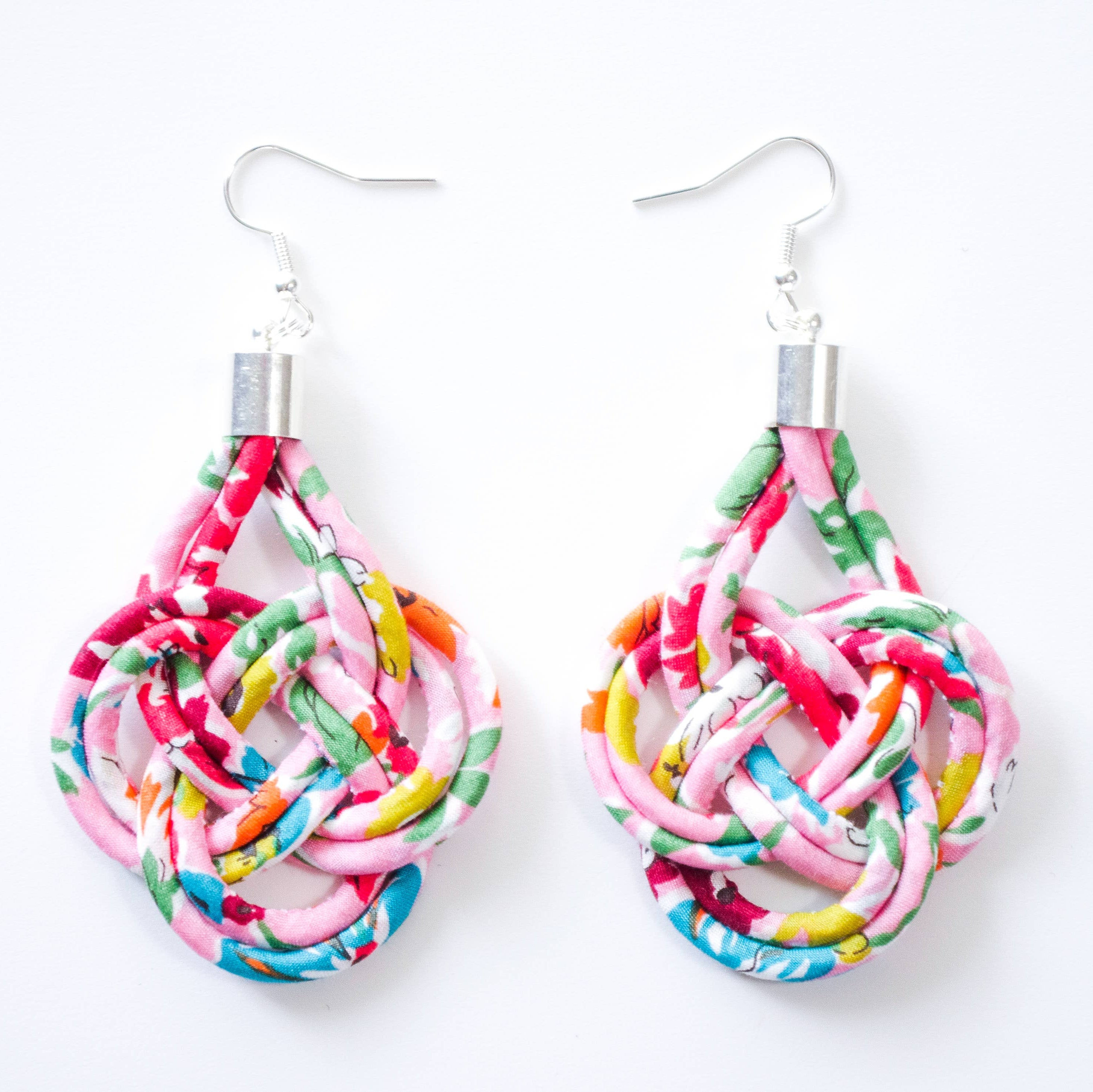 A pair of handmade teardrop-shaped dangle earrings made from knotted, twisted, and woven colorful fabric strips in pink, green, orange, yellow, and blue, hanging from silver fishhook ear wires on a plain white background.