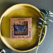 A small square kraft-paper package labeled 'Lily Faith' and 'Mini Pin' sits centered on a round olive-green ceramic plate. The package has a clear window showing a butterfly-shaped pin.