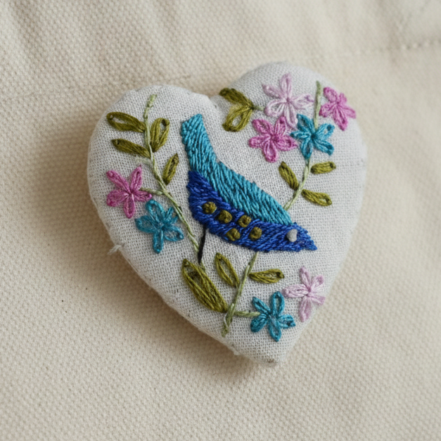 A heart-shaped embroidered patch featuring a blue bird and pink, blue, and green flowers on a white fabric background, pinned to the side of a beige canvas tote bag.