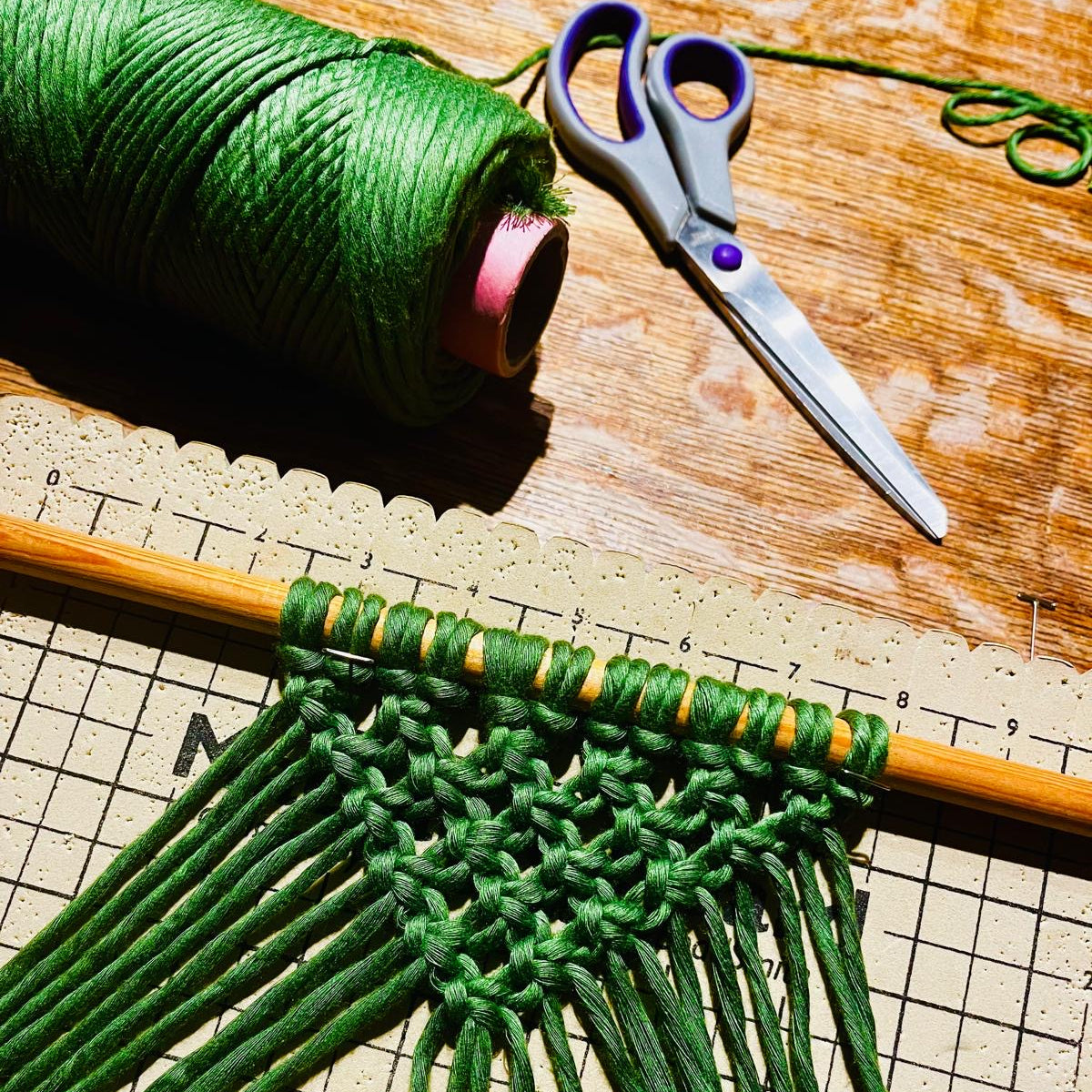 Green yarn with a woven pattern on a wooden surface, accompanied by scissors and a spool of yarn.