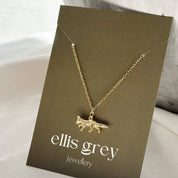 A gold necklace with a small fox pendant is displayed on a dark brown card labeled 'ellis grey Jewellery', set against a soft white fabric background with a textured grey ceramic vase.