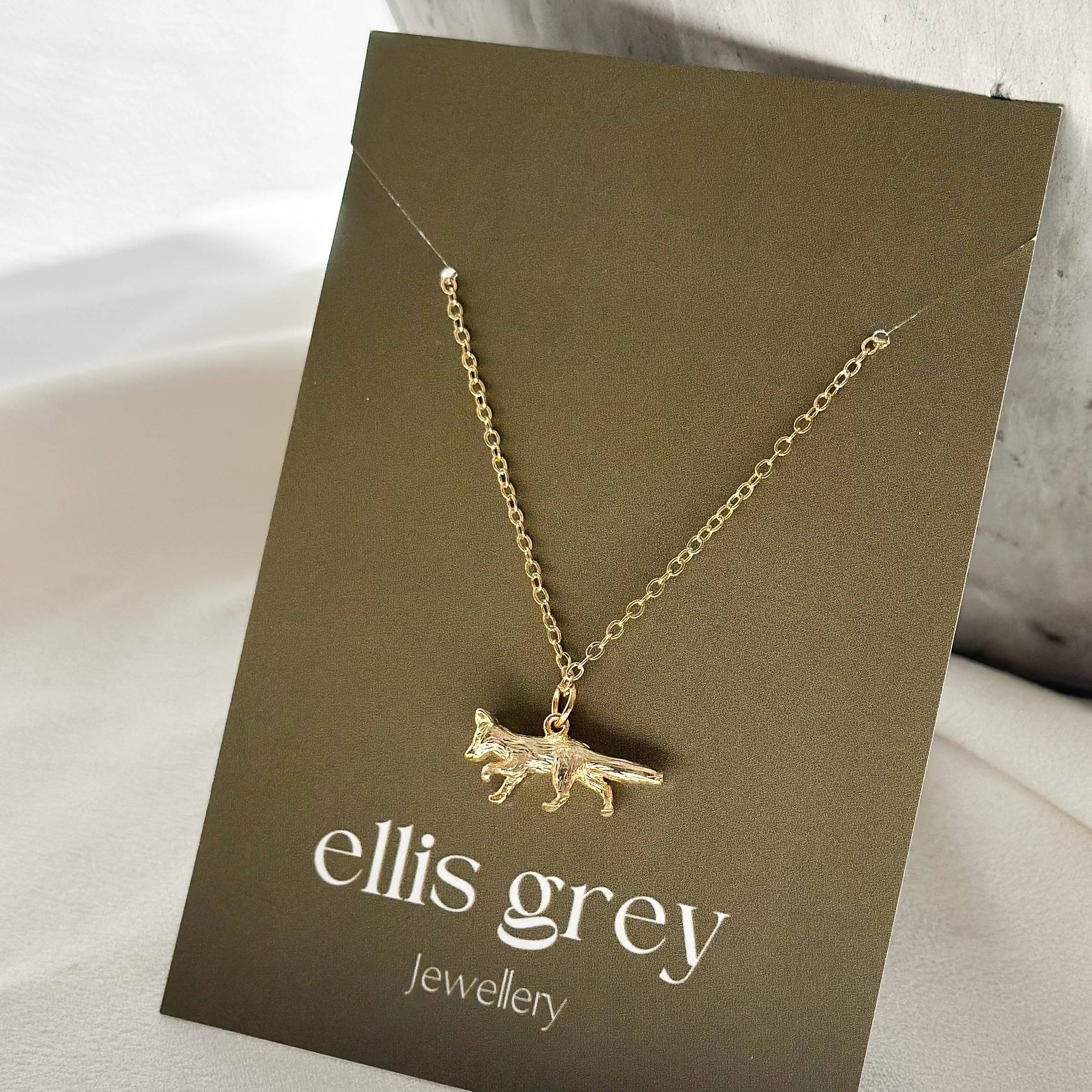 A gold necklace with a small fox pendant is displayed on a dark brown card labeled 'ellis grey Jewellery', set against a soft white fabric background with a textured grey ceramic vase.