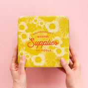 Yellow box with floral design and text held by hands against a pink background