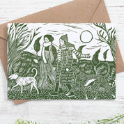 Greeting card with green illustration of two people and a dog in a forest, placed on a white wooden surface with a brown envelope.