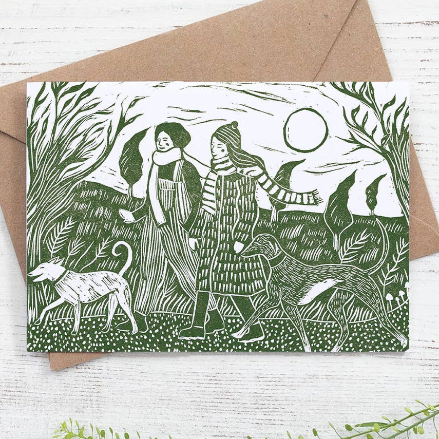Greeting card with green illustration of two people and a dog in a forest, placed on a white wooden surface with a brown envelope.