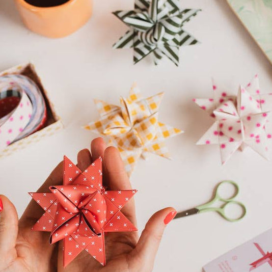 Person holding a red origami star with a crafting setup including scissors, paper, and coffee on a light surface.