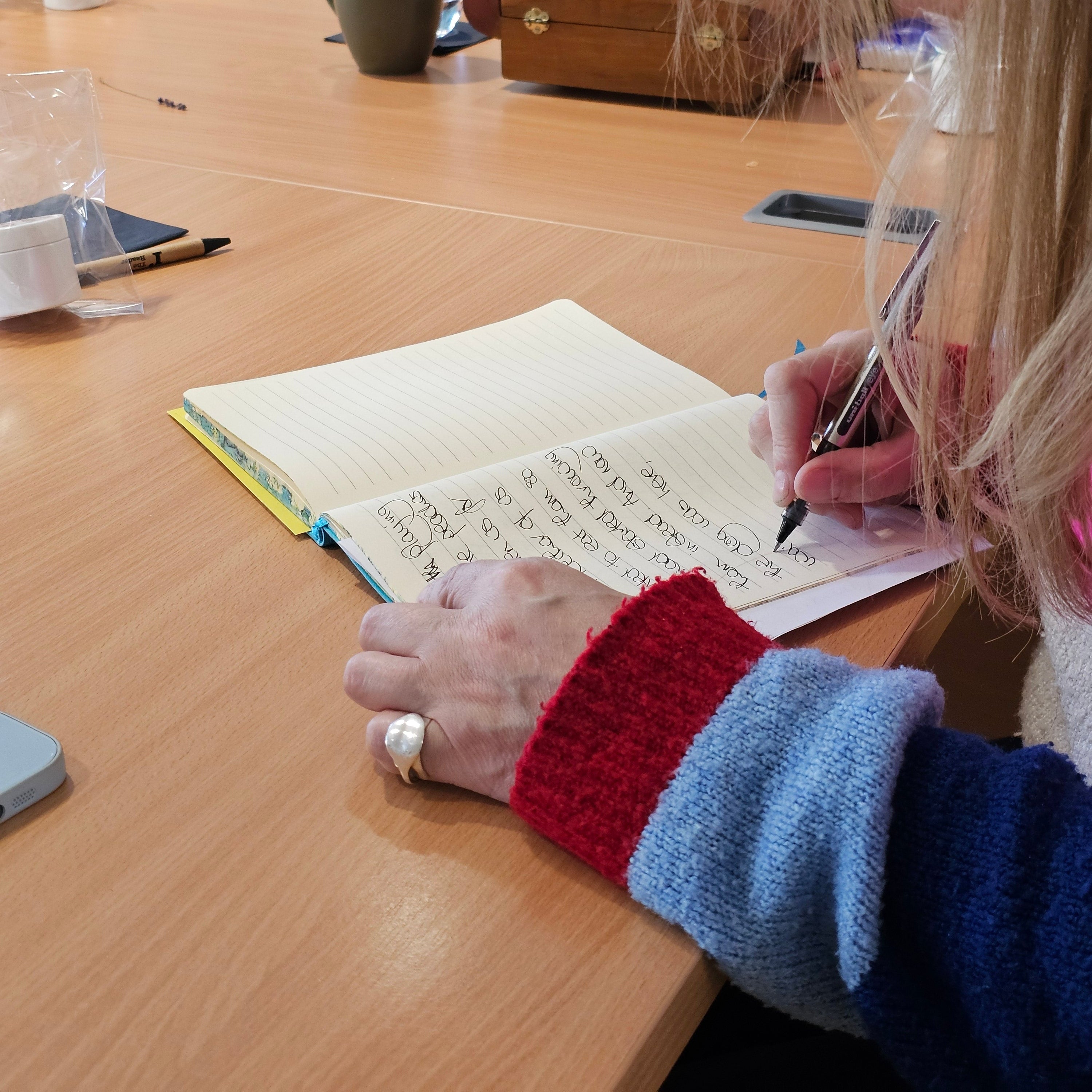 A close-up lifestyle shot of a person writing in a notebook with a black pen, wearing a red, white, and blue striped knit sweater. The scene is set in a warm, sunlit room with a wooden table, other people in the background, and various desk items.