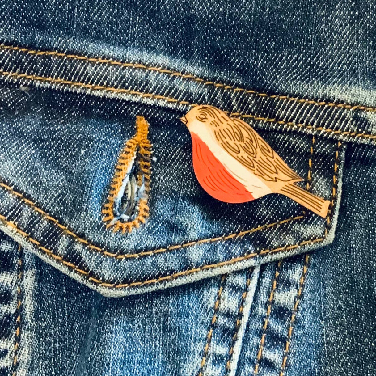 A close-up of a blue denim jacket showing a front pocket with a small bird-shaped enamel pin in gold and orange-red tones, and a gold ring-shaped pin nearby.