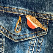 A close-up of a blue denim jacket showing a front pocket with a small bird-shaped enamel pin in gold and orange-red tones, and a gold ring-shaped pin nearby.