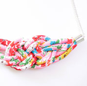 A handmade fabric knot necklace featuring a vibrant pink, red, yellow, and green floral pattern, suspended from a silver chain with silver metal caps.