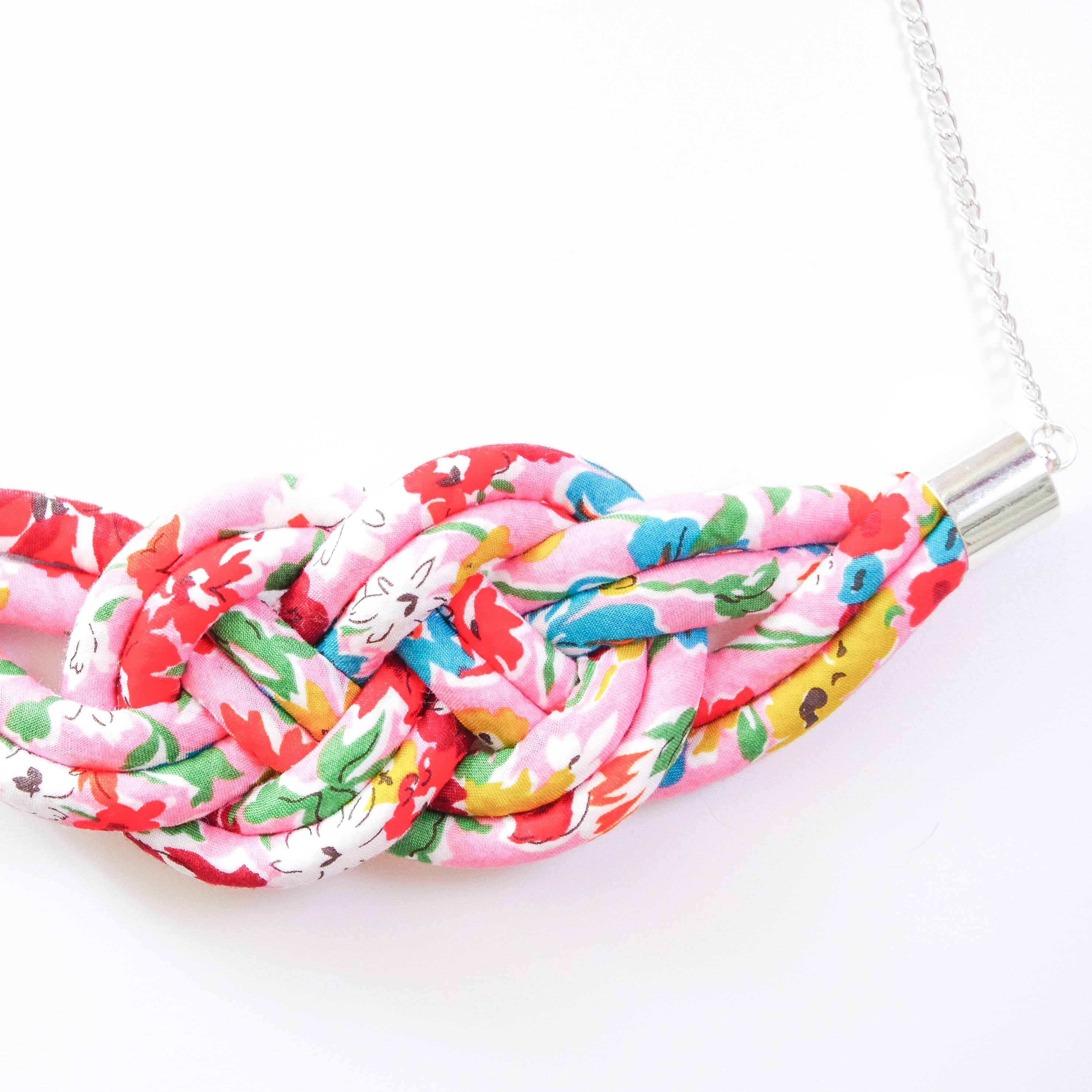 A handmade fabric knot necklace featuring a vibrant pink, red, yellow, and green floral pattern, suspended from a silver chain with silver metal caps.