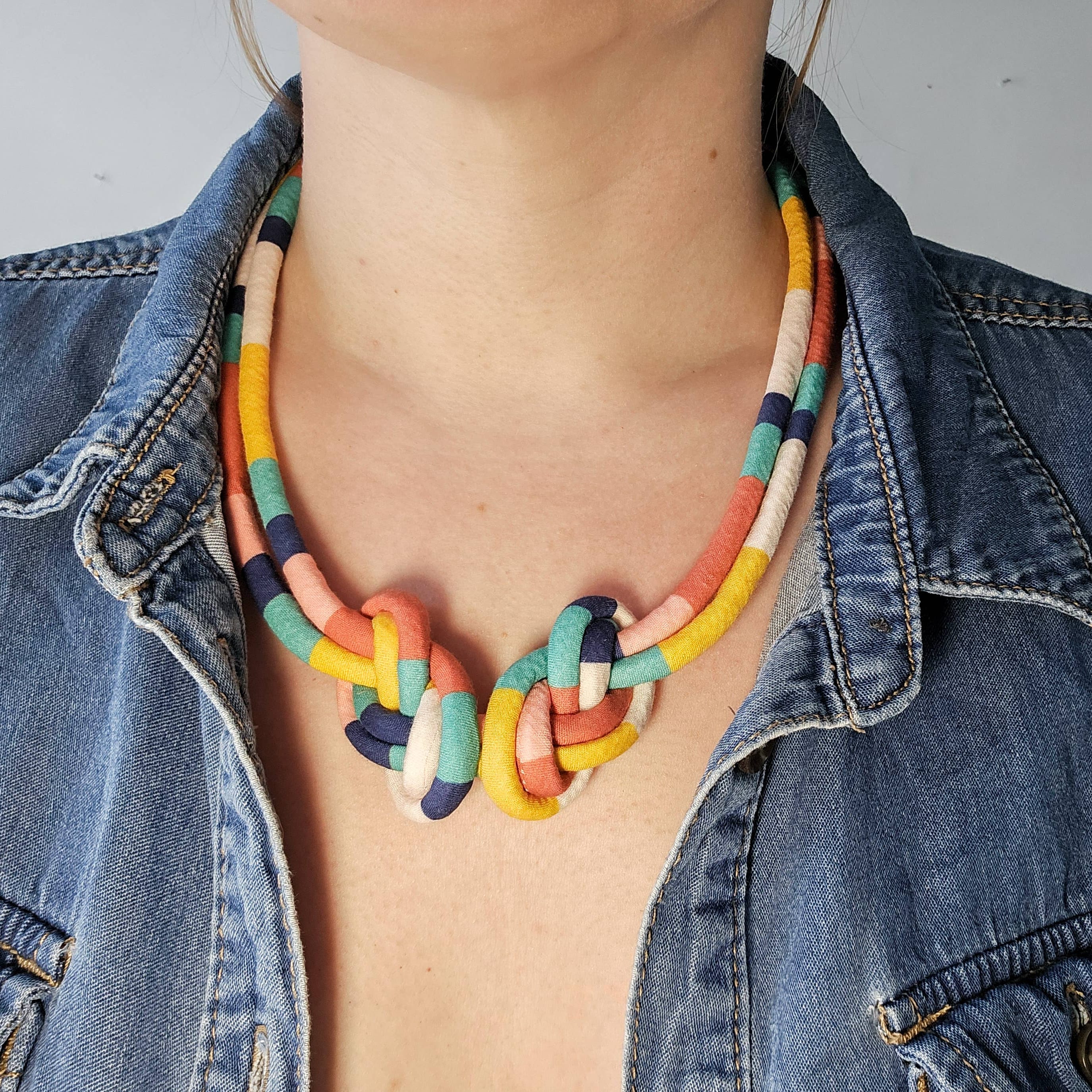 A close-up of a person wearing a colorful braided rope necklace with a knot design, styled with a blue denim jacket.