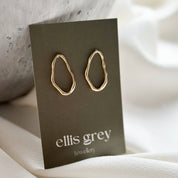 A pair of gold abstract loop hoop earrings displayed on a dark grey card labeled 'ellis grey jewelry', set against a soft cream fabric and a textured grey stone prop.