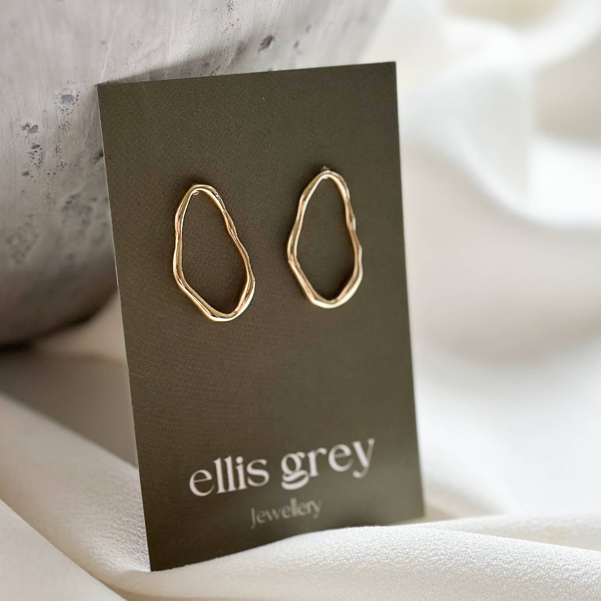 A pair of gold abstract loop hoop earrings displayed on a dark grey card labeled 'ellis grey jewelry', set against a soft cream fabric and a textured grey stone prop.
