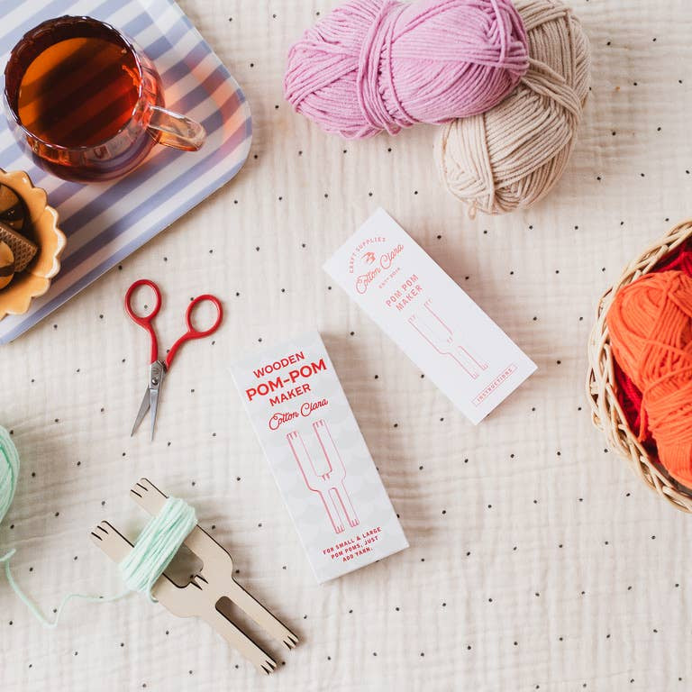 Yarn, scissors, and a pom-pom maker on a textured surface with a cup of tea.