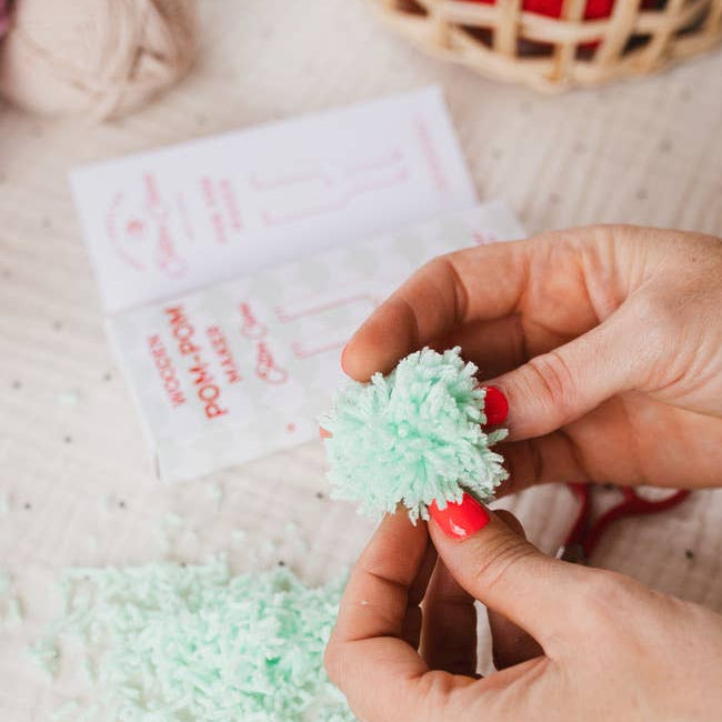 Person making a pom-pom with yarn and a needle on a light surface.