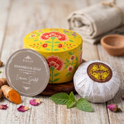 A round tin of DALIT Handmade Cream Secreted soap next to a wrapped soap bar, styled on a rustic wooden table with turmeric roots, rose petals, and a rolled linen cloth.