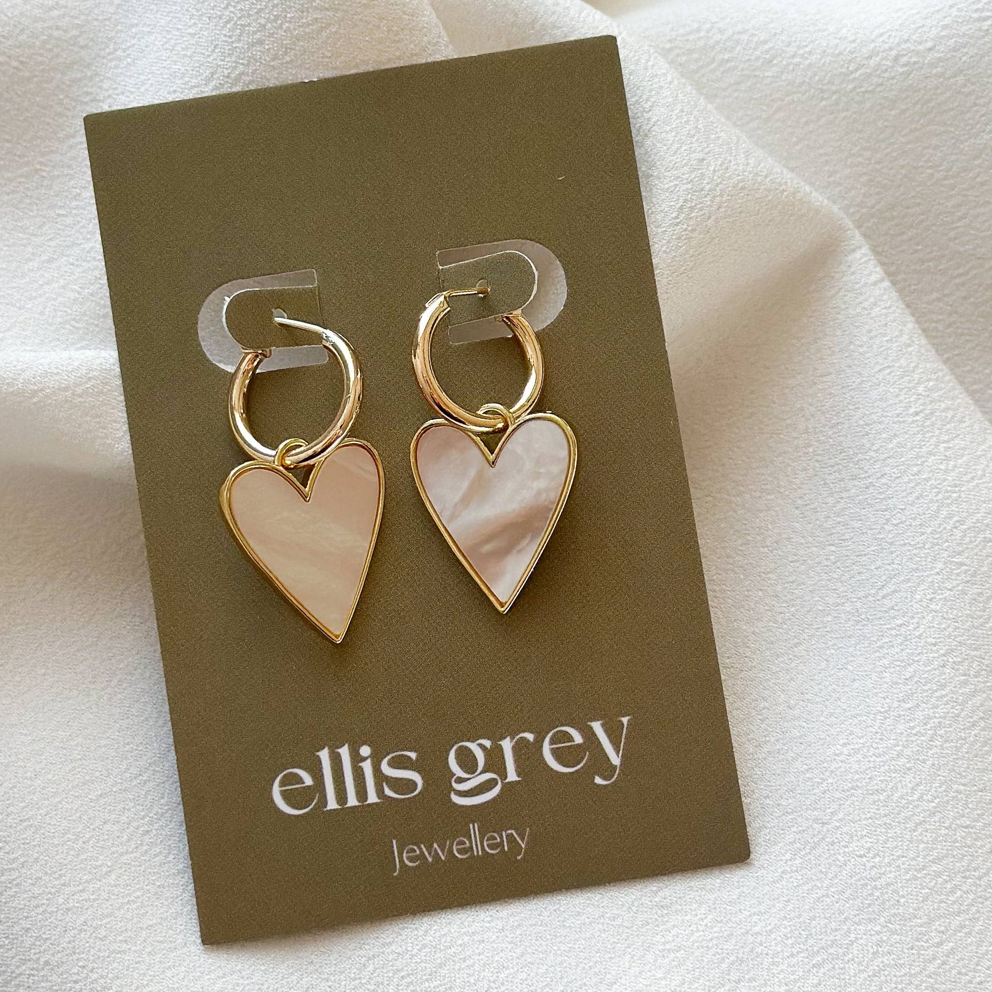 Heart-shaped earrings on an 'ellis grey' jewelry card against a white fabric background
