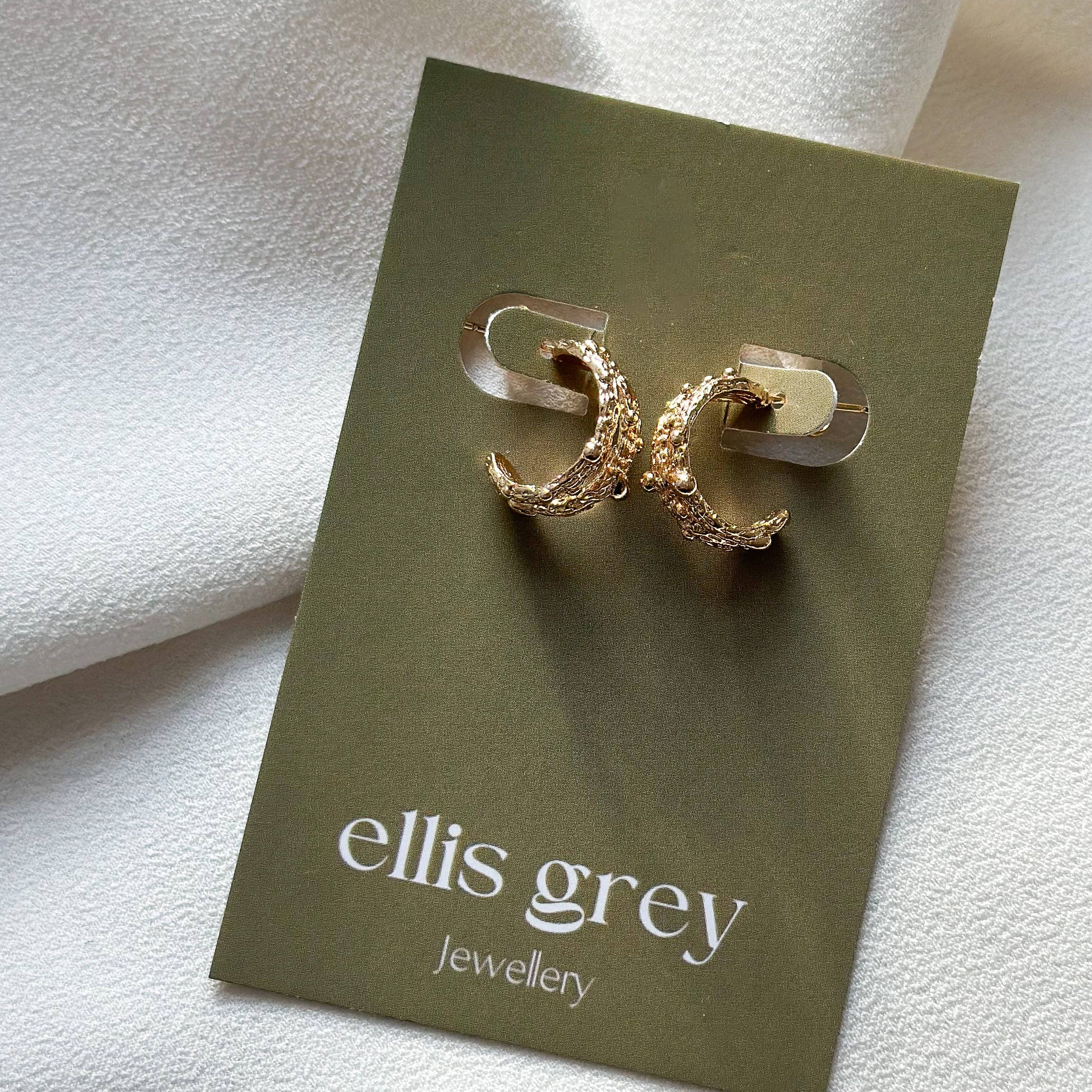Gold earrings on a 'ellis grey' branded card against a white fabric background