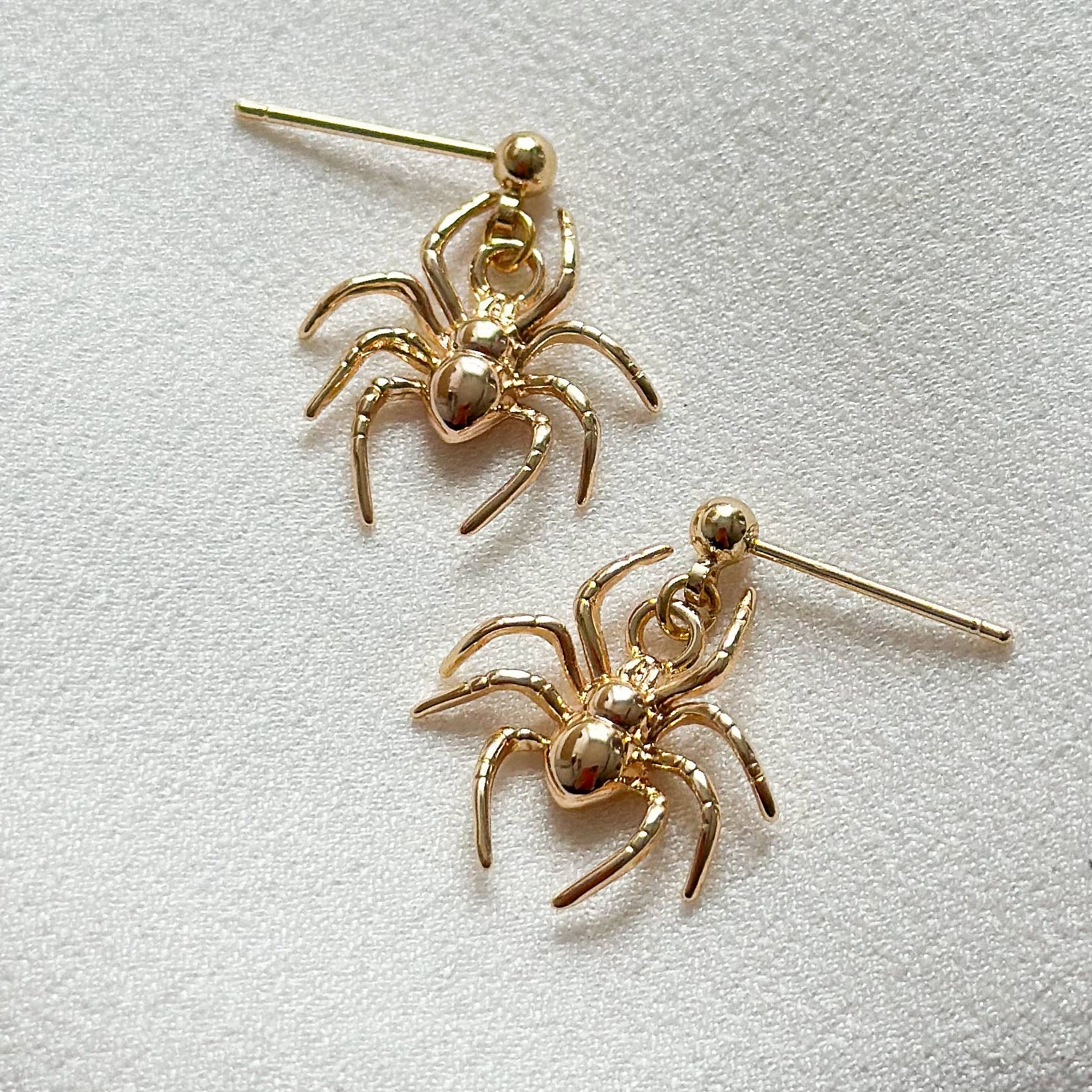 A pair of gold spider-shaped stud earrings resting on a textured white fabric background.