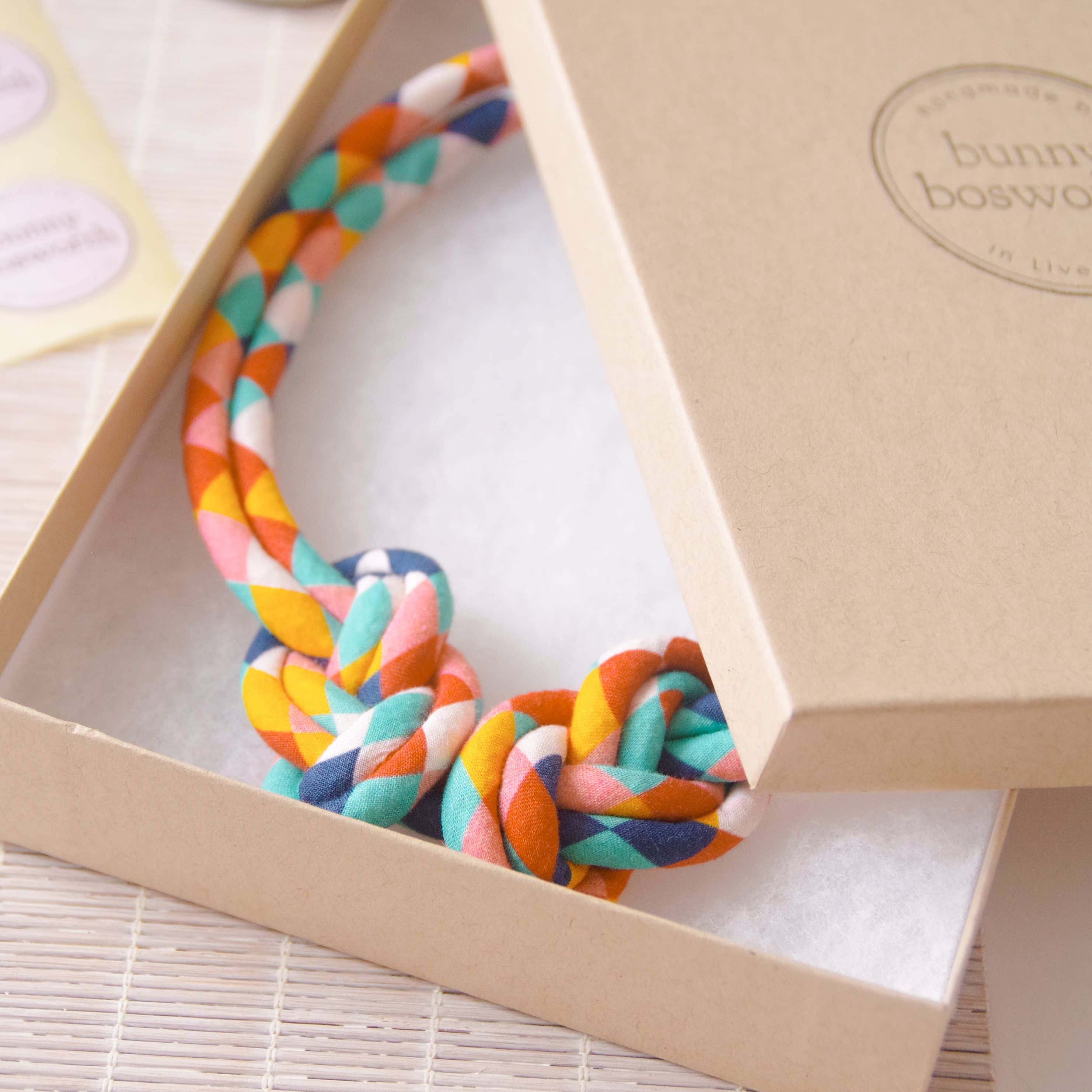 A braided multicolor pet rope toy inside an open kraft paper box, with the branded lid leaning against it. The toy is made of braided rope in orange, teal, pink, yellow, and blue, and the box has a visible bunny bosworth logo.