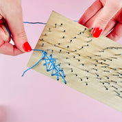 A close-up of hands with red nail polish threading blue string onto a wooden nail art board against a pink background.