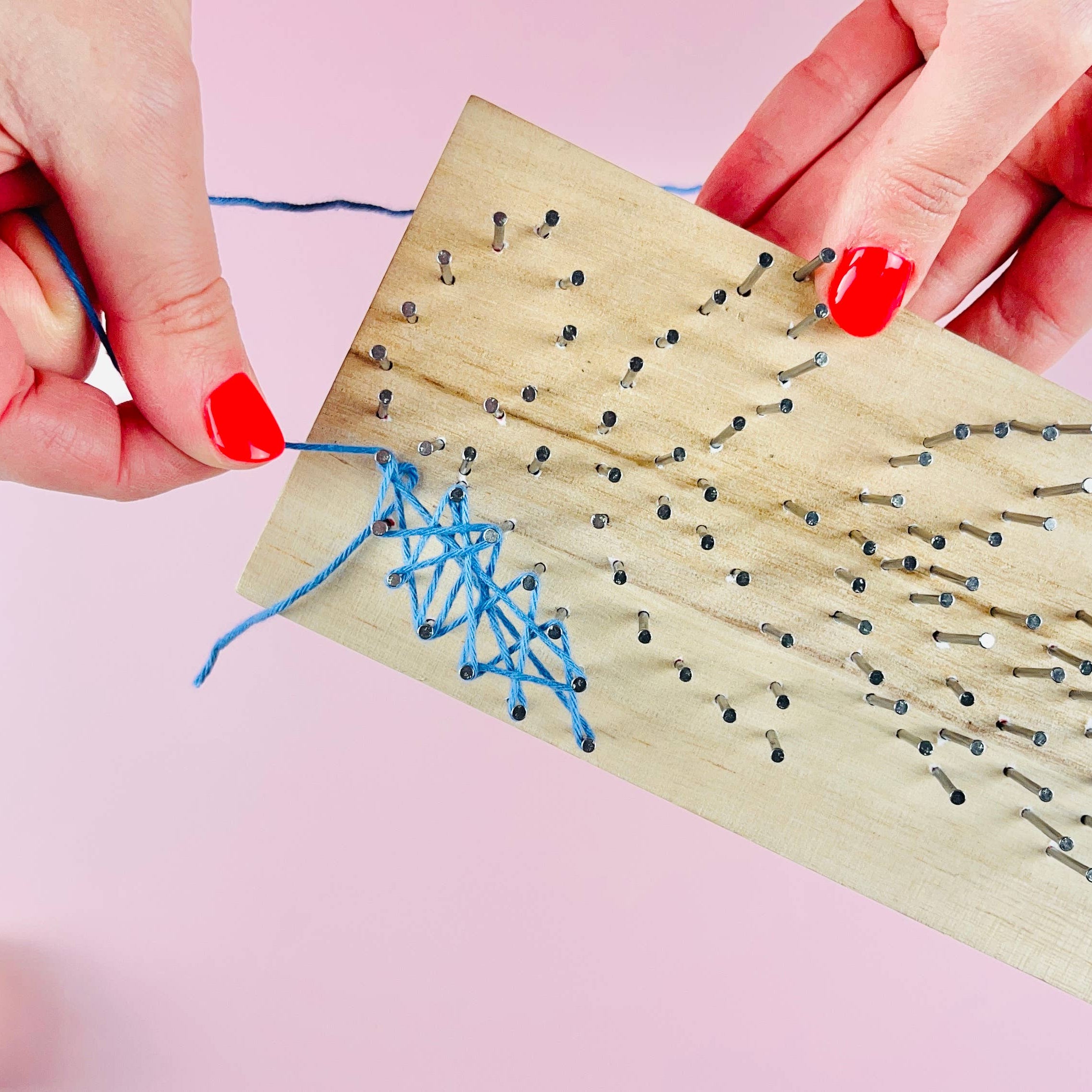 A close-up of hands with red nail polish threading blue string onto a wooden nail art board against a pink background.