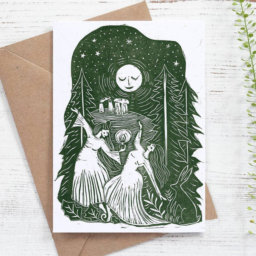 Greeting card with green and white illustration of two women in a forest setting, accompanied by a brown envelope on a light wooden surface.