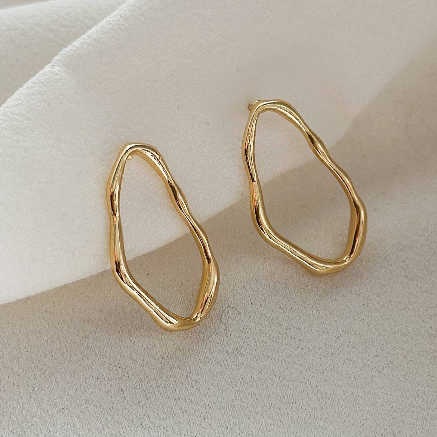 A pair of gold abstract wavy hoop earrings displayed on a soft, draped white fabric background.