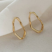 A pair of gold abstract wavy hoop earrings displayed on a soft, draped white fabric background.