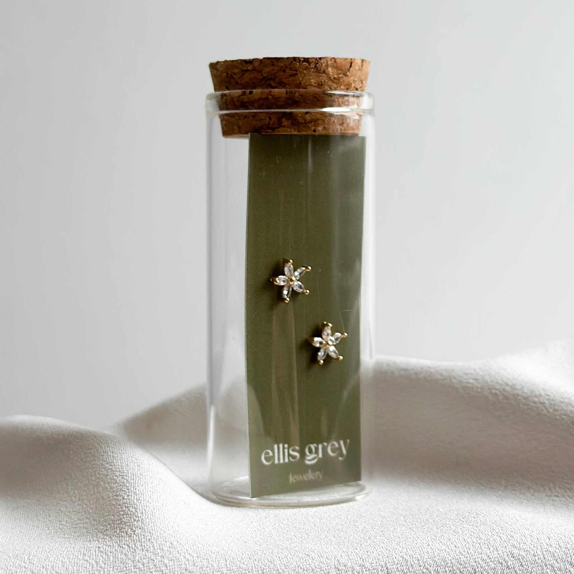 A pair of small star-shaped stud earrings in silver-toned metal, displayed on a dark olive green card inside a clear glass tube with a cork stopper. The card is branded with 'ellis grey' and is set against a draped white fabric backdrop.