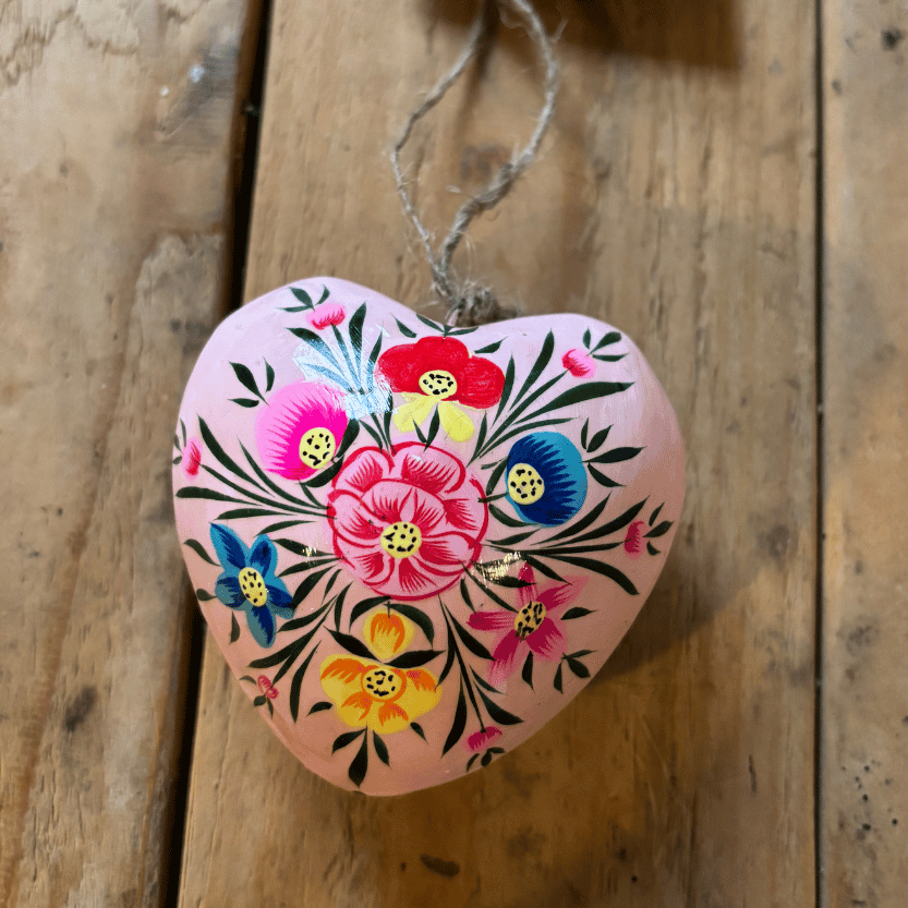 A heart-shaped wooden ornament with a hand-painted floral design in pink, red, yellow, and blue, hanging from a jute string with a 'DALIT' tag, set against a rustic wooden plank background.