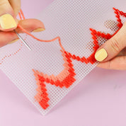 A close-up of hands with pale yellow nail polish stitching a red and orange gradient pattern onto a white mesh counting board with a needle.