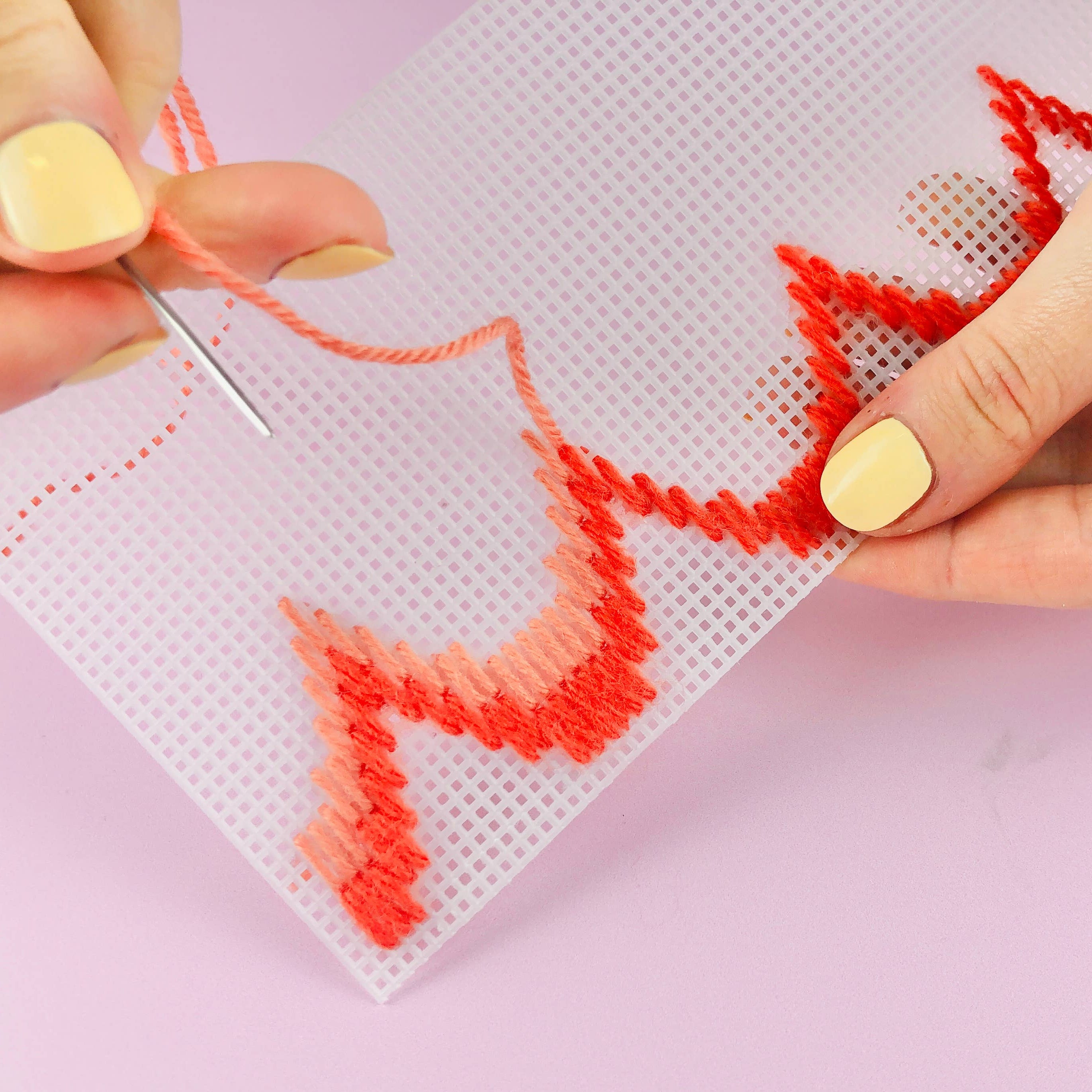 A close-up of hands with pale yellow nail polish stitching a red and orange gradient pattern onto a white mesh counting board with a needle.