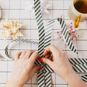 Person wrapping a gift with striped ribbon on a checkered tablecloth.